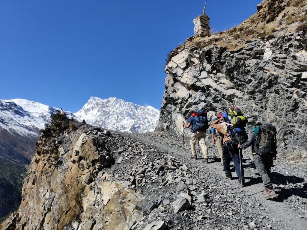 Sherpas, porters and trekkers climbing steep loose trail with snow capped mountains in the distance on the Annapurna Circuit