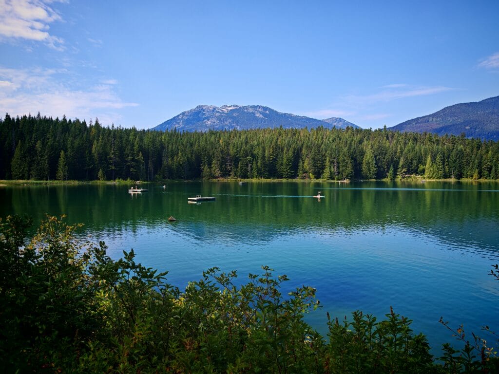 The deep blue waters of Lost Lake in Whistler with a fine forest surrouding the lake with swimmers and floating pontoons and a mountainous backdrop