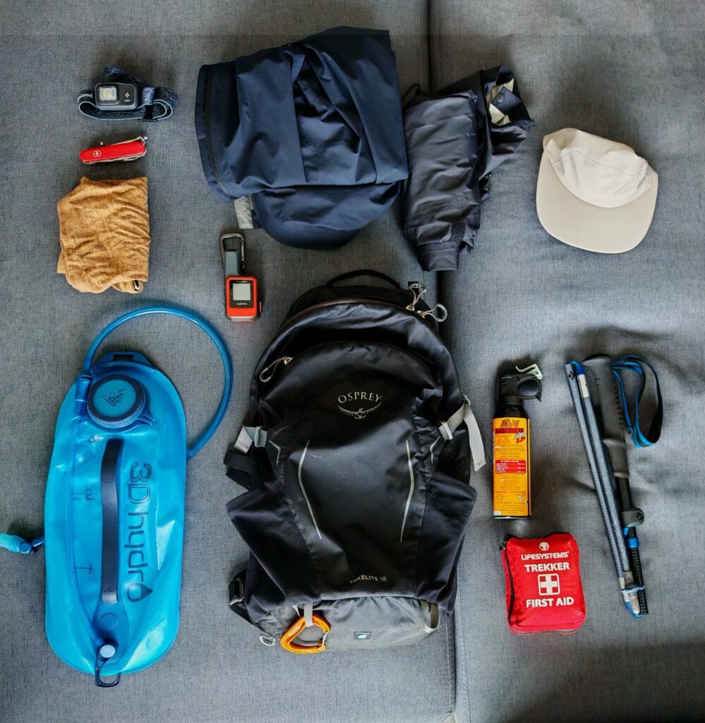 A flat lay out of essential hiking gear list for Canada, including backpack, bear spray, poles, premium waterproof, hydration bladder, cap, headtorch and first aid kit on a fabric background.