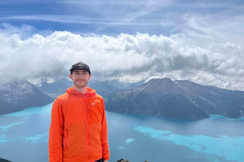 Male hiker wearing an orange Arc'teryx Proton LT hoody standing on a rocky mountain summit above a bright turquoise glacial lake in Canada.