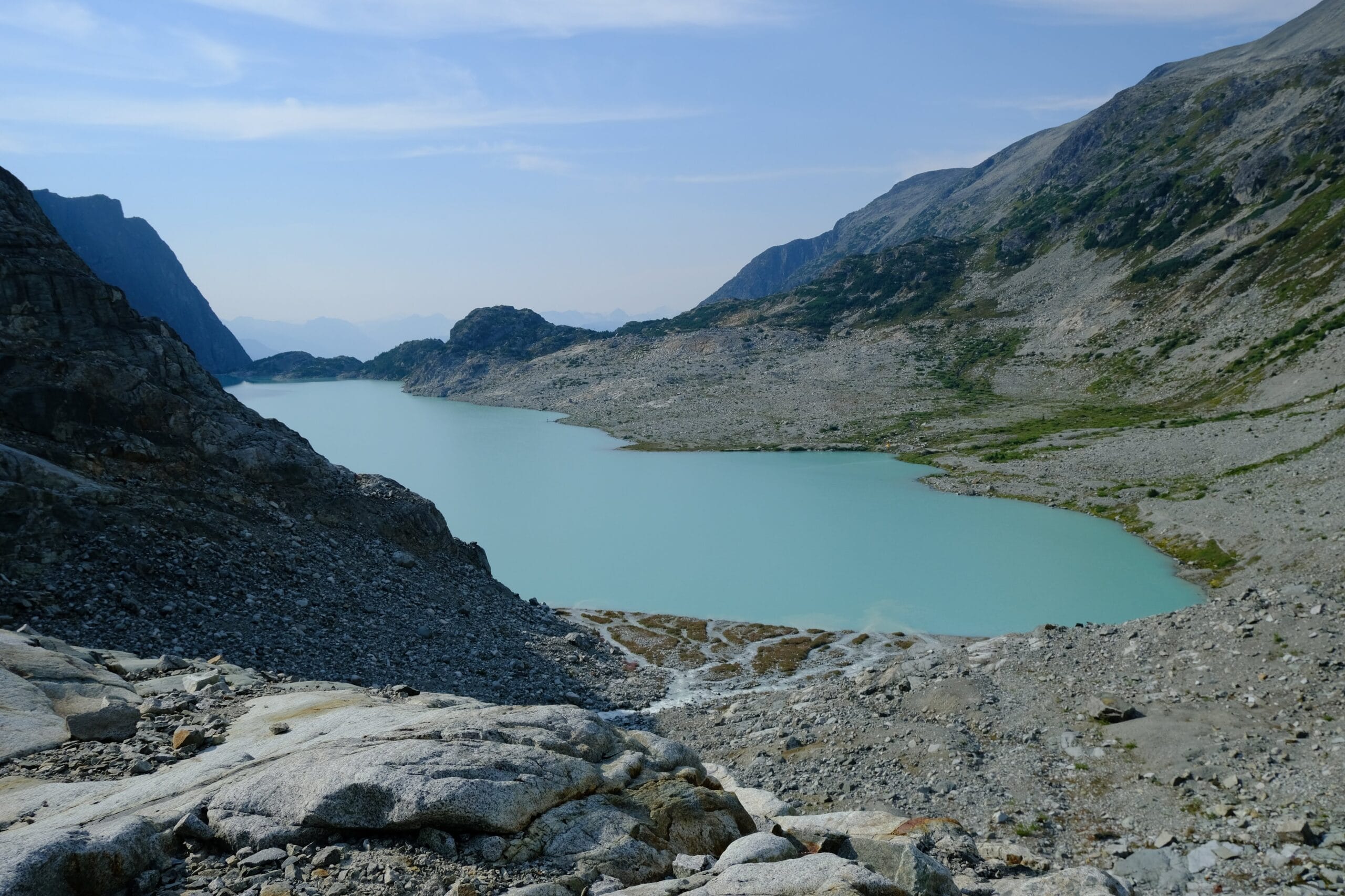 Wedgemount Lake with turquoise waters next to the alpine mountain slopes with blue skies above and a rocky outcrop in the foreground