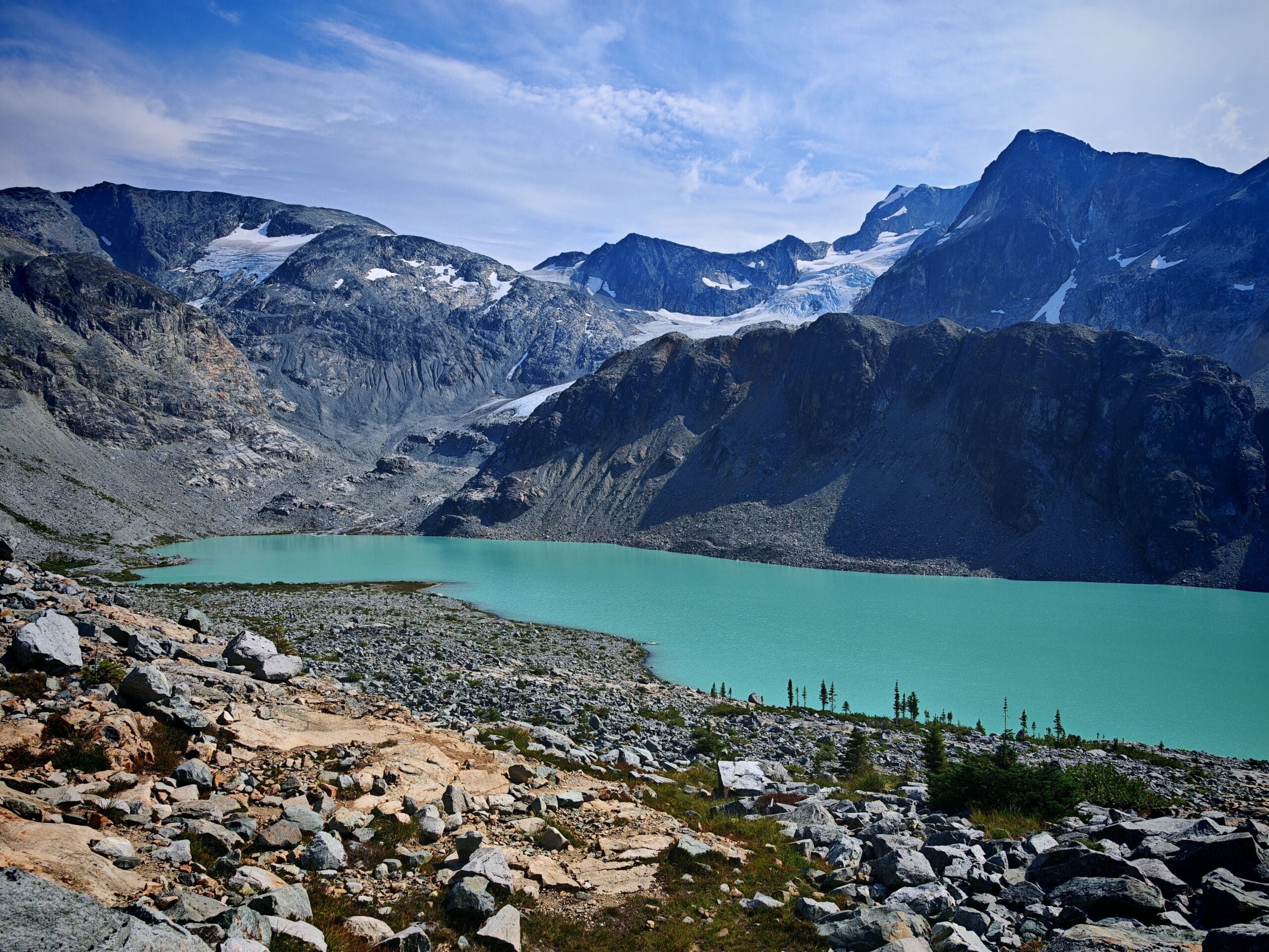 The turquoise waters of Wedgemount Lakes sits below rocky mountains surrounding the lake and with a rocky path seen in the foreground