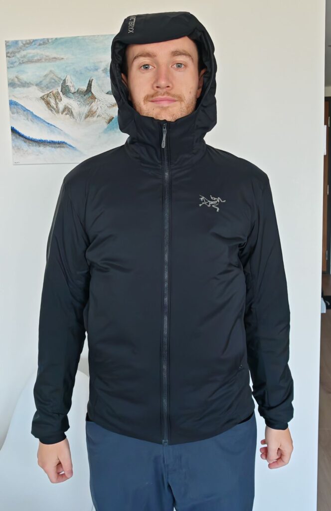 A man stood wearing the Arc'teryx Atom Hoody in Black size Small Men's against a white wall with the stormhood up showing the Arc'teryx brand text